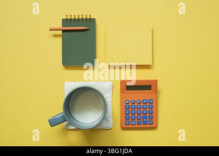 Bloc-notes vert, papier mémo, crayon de couleur, tasse de lait, calculatrice isolée sur fond jaune. flat lay, vue de dessus, espace de copie Banque D'Images