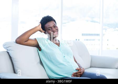 Femme afro-américaine assise sur un canapé gris berçant le ventre dans un salon moderne avec de grandes fenêtres Banque D'Images