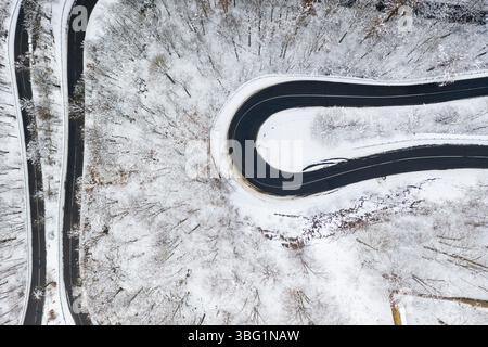 Vue aérienne par drone d'une route en asphalte noir sinueuse serpentant à travers un paysage forestier enneigé d'hiver Banque D'Images