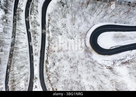 Vue aérienne par drone d'une route en asphalte noir sinueuse serpentant à travers un paysage forestier enneigé d'hiver Banque D'Images