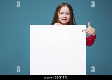 Isolé sur bleu, magnifique enfant brune caucasien en chemise à carreaux rouge et Jean bleu avec de longs cheveux tenir l'affiche vide, pointez vers l'affiche, regardez Banque D'Images