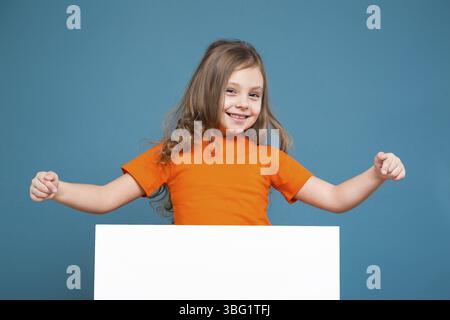 Isolé sur bleu, bel enfant caucasien avec de longs cheveux, en t-shirt orange et en Jean bleu montrent une affiche vide, regardez la caméra Banque D'Images
