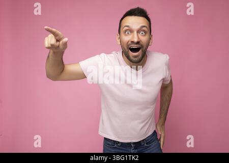 Photo de beau émotionnel heureux positif stupéfait brunet homme dans un t-shirt rose décontracté isolé sur fond rose avec un espace de copie pour le texte et la crier Banque D'Images