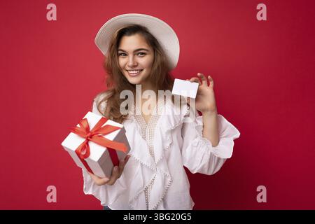 Jeune belle européenne élégante brune femme portant un chemisier blanc et chapeau à la mode isolé sur fond rouge avec des émotions sincères positives. Banque D'Images