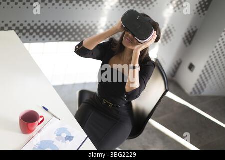 Beauté jeune brunette afroamerican femme d'affaires en noir costume d'affaires, lunettes et montre coûteuse s'asseoir à la table dans le bureau, coupe rouge sur la table Banque D'Images