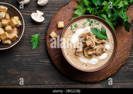 Soupe à la crème de champignons dans un bol garni de croûtons, champignons frais et herbes sur fond de bois foncé, vue de dessus, espace copie. Repas saisonnier d'automne - Banque D'Images