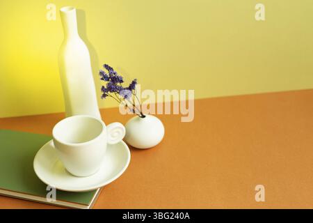 Carnet, tasse à café blanche, vase de fleur pourpre sèche sur table orange. Fond de mur jaune Banque D'Images