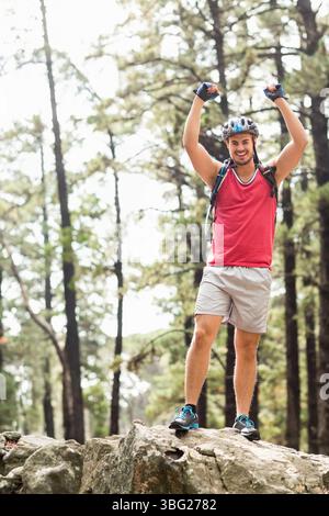 Homme debout au sommet du rocher dans la forêt de pins levant les bras portant casque de vélo et sac à dos d'hydratation Banque D'Images