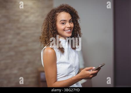 Femme afro-américaine en blouse blanche debout tout en souriant, tenant smartphone dans le couloir du bureau Banque D'Images