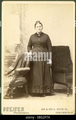 Cette photographie, prise par John A. Pfeifer, montre une femme à la coiffure chignon haute, portant un col en dentelle et une robe noire avec un ourlet de jupe décoré. L'image reflète la mode de la fin du XIXe siècle. Banque D'Images