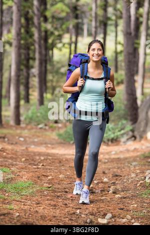 Randonneur féminin à la fin de la vingtaine marchant le long de la piste des conifères portant des vêtements de sport et portant un sac à dos Banque D'Images