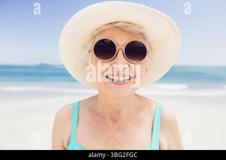 Femme senior souriant sur la plage de sable blanc portant chapeau de paille ronde lunettes de soleil maillot de bain turquoise Banque D'Images