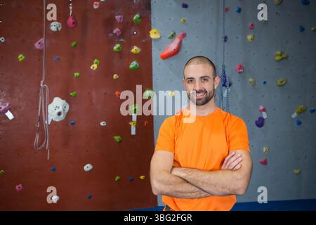 Homme debout bras croisés portant chemise orange à la salle de gym d'escalade avec corde et prises, espace de copie Banque D'Images