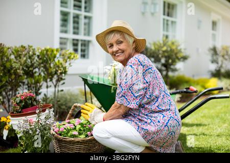 Femme senior agenouillée sur la cour arrangeant des pots de fleurs près de brouette portant chapeau de paille, espace de copie Banque D'Images