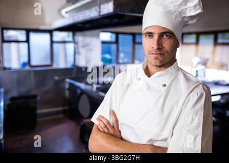 Chef masculin debout avec les bras croisés sous le capot dans la cuisine du restaurant portant un manteau blanc et un chapeau Banque D'Images