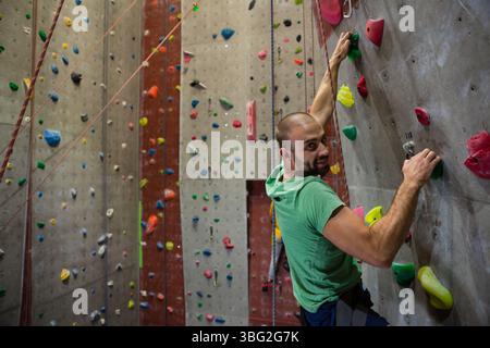 Homme grimpant mur de roche artificielle dans la salle de gym saisissant des prises multicolores avec harnais de corde, espace de copie Banque D'Images