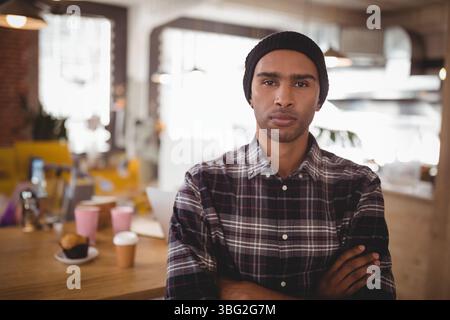 Homme afro-américain debout avec les bras croisés à la table du café avec ordinateur portable, tasses à café et muffin Banque D'Images