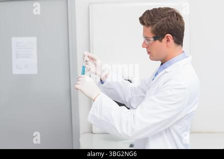 Un scientifique masculin portant une blouse de laboratoire et des lunettes inspecte la solution bleue dans un tube à essai au banc Banque D'Images