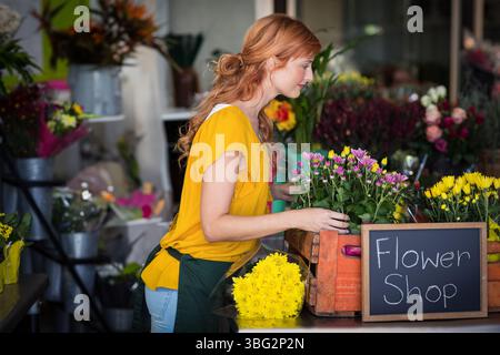 Fleuriste féminine arrangeant les mamans jaunes, marguerites sur le comptoir en bois avec signe de tableau noir dans le magasin de fleurs Banque D'Images