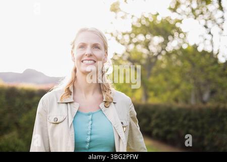 Femme dans ses années 30 souriant et regardant vers le haut tout en se tenant debout dans le jardin ensoleillé portant une veste beige Banque D'Images