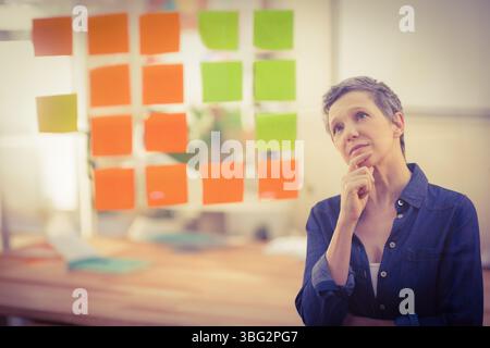 Femme senior debout au panneau de verre avec des notes adhésives orange et vertes dans le bureau, espace de copie Banque D'Images