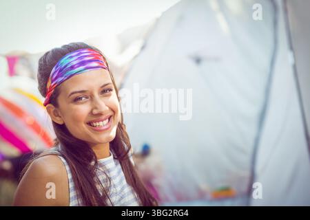 Femme souriant à la caméra debout parmi les tentes de festival de toile dans le serre-tête tie-dye, débardeur rayé Banque D'Images