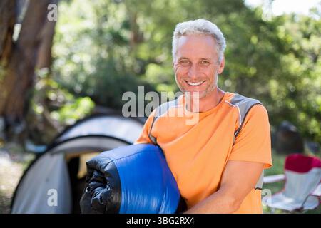Homme d'âge moyen portant un T-shirt orange portant un sac de couchage bleu au camping sous tente et chaise rouge Banque D'Images
