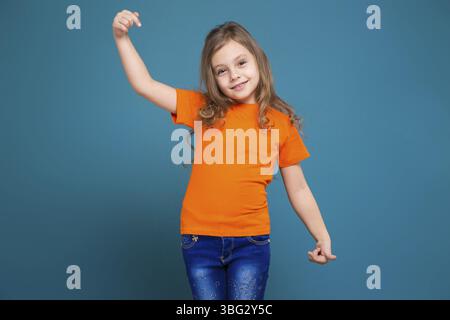 Isolé sur bleu, bel enfant caucasien avec de longs cheveux, en t-shirt orange et en Jean bleu, pointe vers elle-même, main haute Banque D'Images