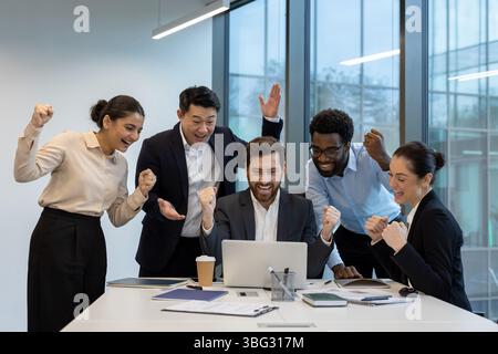 Une équipe d'affaires diversifiée célèbre un succès, regardant un ordinateur portable dans un environnement de bureau moderne. Leurs expressions montrent le bonheur et l'accomplissement. Banque D'Images