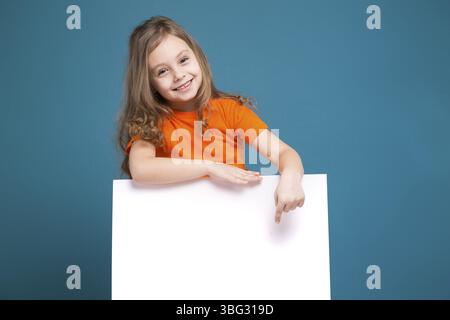 Isolé sur bleu, bel enfant caucasien avec de longs cheveux, en t-shirt orange et en Jean bleu montrent une affiche vide, se fie à l'affiche, pointez sur le po Banque D'Images