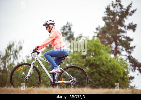 Femme chevauchant le vélo de montagne blanc jusqu'à la colline herbeuse au bord de la forêt portant casque et lunettes de soleil Banque D'Images