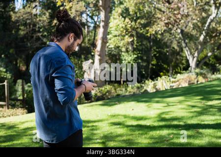 Photographe masculin ajustant l'appareil photo reflex numérique sur la pelouse d'herbe dans le parc avec le filtrage de la lumière du soleil à travers les arbres Banque D'Images