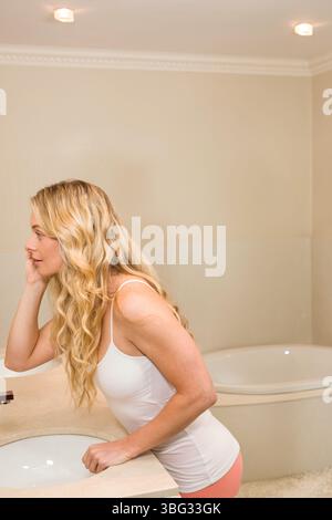 Femme blonde portant un débardeur blanc et un short rose s'appuyant sur le comptoir de l'évier, regardant la baignoire Banque D'Images