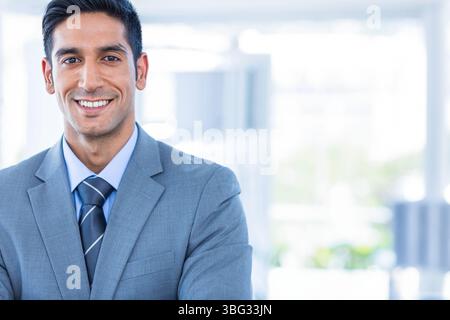 Homme debout avec les bras croisés dans un espace de travail moderne lumineux, vêtu d'un costume gris et d'une cravate rayée Banque D'Images
