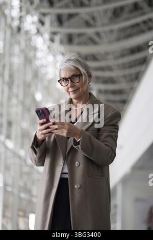 Une femme d'affaires âgée se tient confiante dans un cadre de bureau contemporain, s'engageant avec son smartphone tout en vêtue d'un manteau élégant. Ses expres Banque D'Images