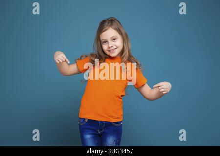 Isolé sur bleu, bel enfant caucasien aux cheveux longs, en t-shirt orange et en Jean bleu Banque D'Images
