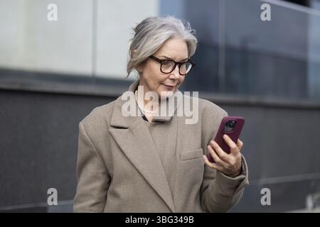 Une femme d'affaires âgée, vêtue d'un manteau élégant, est concentrée sur son smartphone alors qu'elle se tient près d'un immeuble de bureaux contemporain. Elle semble engag Banque D'Images