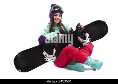 Isolé sur blanc, jolie fille caucasienne en tenue de ski violette, pantalon rouge, bottes de neige bleues et chapeau de laine violette assis jambes croisées avec neige noire Banque D'Images