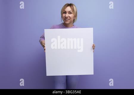 Attraktive blonde frau mit einem blatt papier mit einem mackup fuer eine werbebotschaft auf einem lila hintergrund Banque D'Images