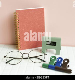Bureau table avec carnet, lunettes, horloge, clip, fournitures sur bureau blanc. Fond de mur rose Banque D'Images