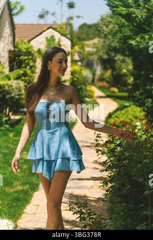 Une femme brune des années 20 dans une robe bleue marche gracieusement dans un jardin d'hôtel de luxe pendant la journée. Banque D'Images