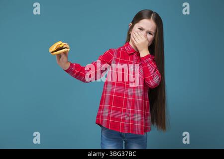 Isolé sur bleu, beau caucasien brunette enfant dans la chemise à carreaux rouge et Jean bleu avec les cheveux longs tenir hamburger, malheureux, dégoût, ferme son no Banque D'Images