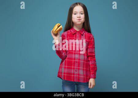 Isolé sur bleu, beau caucasien brunette enfant dans la chemise à carreaux rouge et Jean bleu avec les cheveux longs tenir hamburger, manger, bouche pleine Banque D'Images