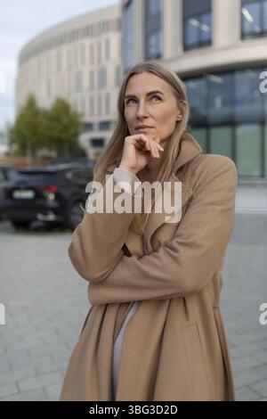 Une femme vêtue d'un manteau bronzé se tient devant un centre d'affaires moderne. Elle a la main sur le menton, levant les yeux avec soin Banque D'Images