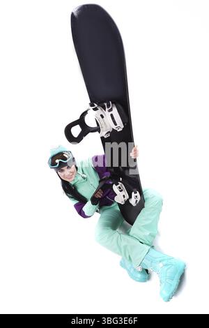 Image verticale, isolée sur blanc, jolie femme caucasienne en tenue de ski violette, lunettes de ski bleues et pantalon bleu assis jambes croisées et tenir bla Banque D'Images