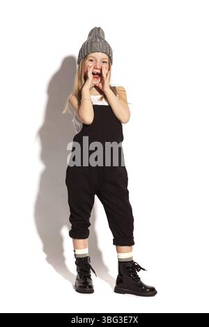 Isolé sur blanc, joyeuse fille aux cheveux rouges sans dent en salopette noire, t-shirt blanc, chapeau gris et chaussures laquées à lacets, se tenant les mains près Banque D'Images