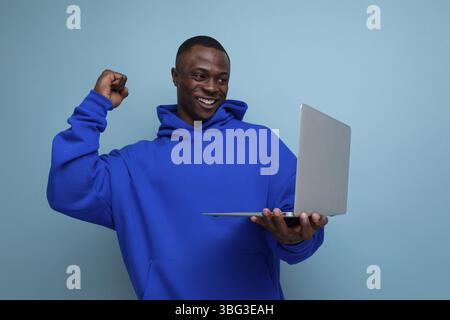 Beau pigiste américain de 25 ans à peau foncée dans un sweat à capuche élégant avec ordinateur portable sur fond avec espace de copie Banque D'Images