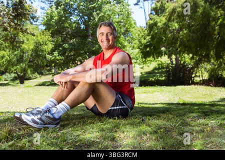 Homme senior assis avec les genoux pliés sur l'herbe ensoleillée dans le parc souriant portant des chaussures de course grises Banque D'Images