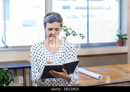 Femme d'affaires écrivant dans un cahier à l'aide d'un stylo près d'un plan roulé aux fenêtres de bureau lumineuses Banque D'Images