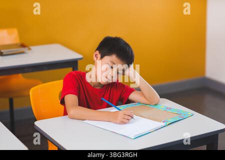 Garçon asiatique reposant la tête sur l'écriture à la main dans un cahier de travail coloré au bureau de l'école avec un crayon Banque D'Images